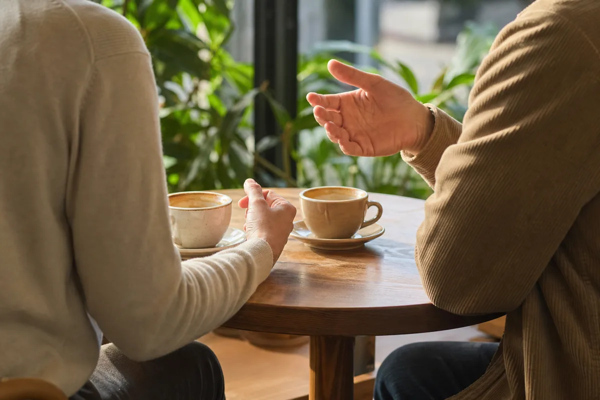 Deux personnes cadrées de dos assises face à face dans un café lumineux, tasses posées sur la table, plantes vertes floues en arrière-plan