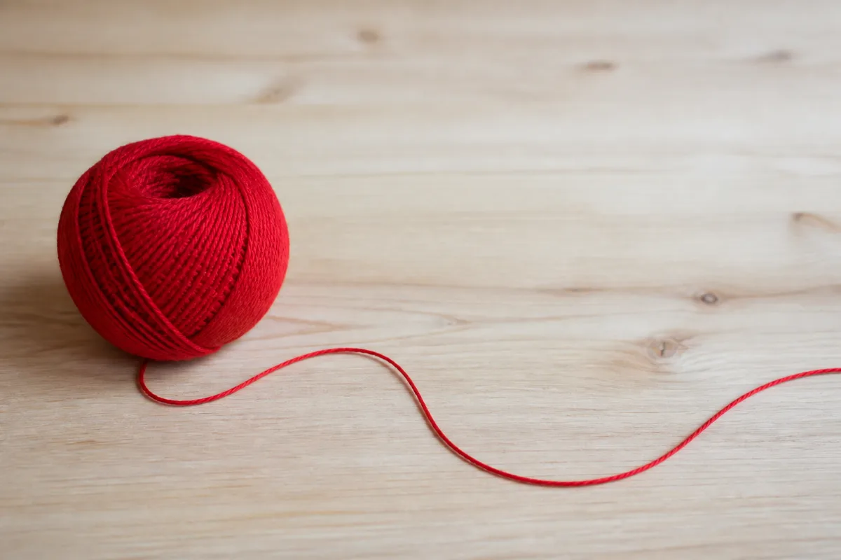 Pelote de fil de coton rouge dont le fil se déroule en longue courbe sur une table en bois clair, plan rapproché, lumière naturelle douce